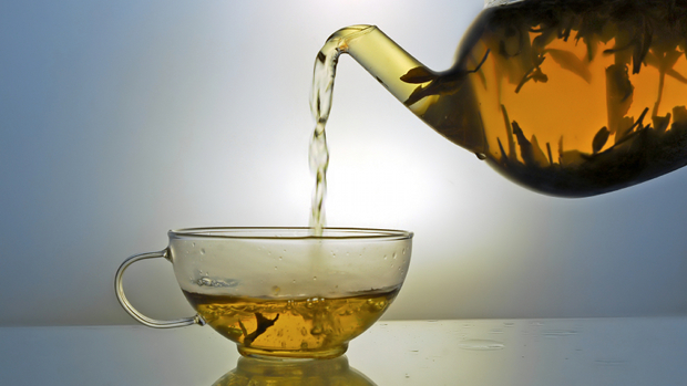 Green tea pouring into glass cup from teapot on blue background